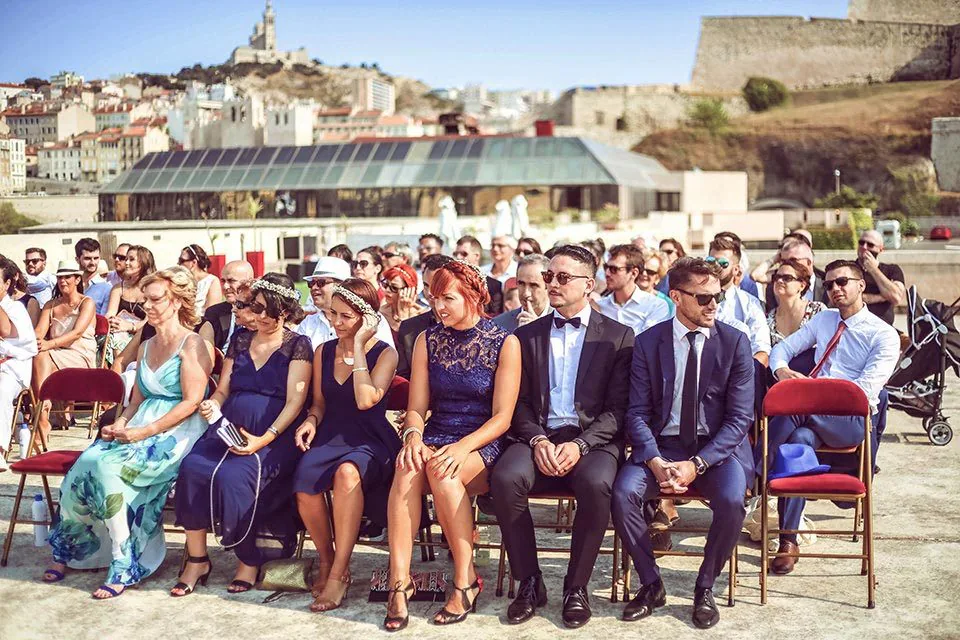 Invités assis lors d’une cérémonie en plein air, Notre-Dame de la Garde en arrière-plan, à Marseille