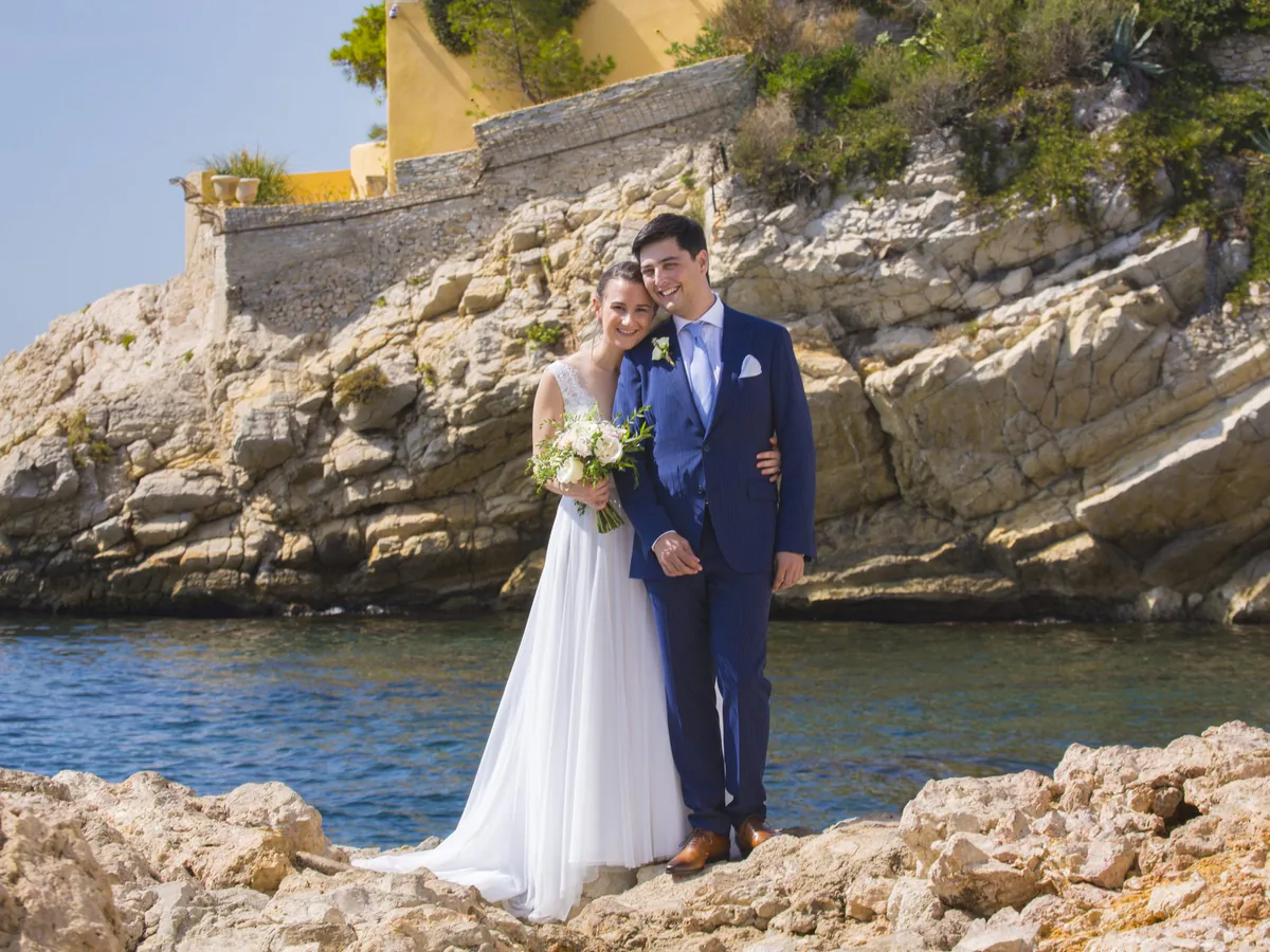 Couple de mariés au bord de l’eau dans une calanque de Marseille, falaise et bastide en arrière-plan
