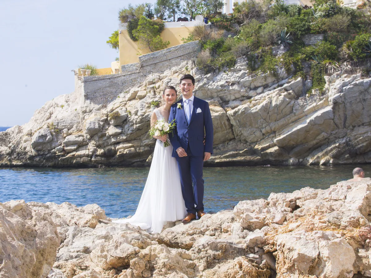 Couple de mariés au bord de l’eau dans une calanque, falaise et bastide en arrière-plan, près de Marseille