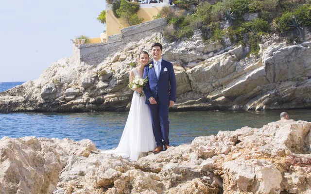 Couple de mariés au bord de l’eau dans une calanque, falaise et bastide en arrière-plan, près de Marseille