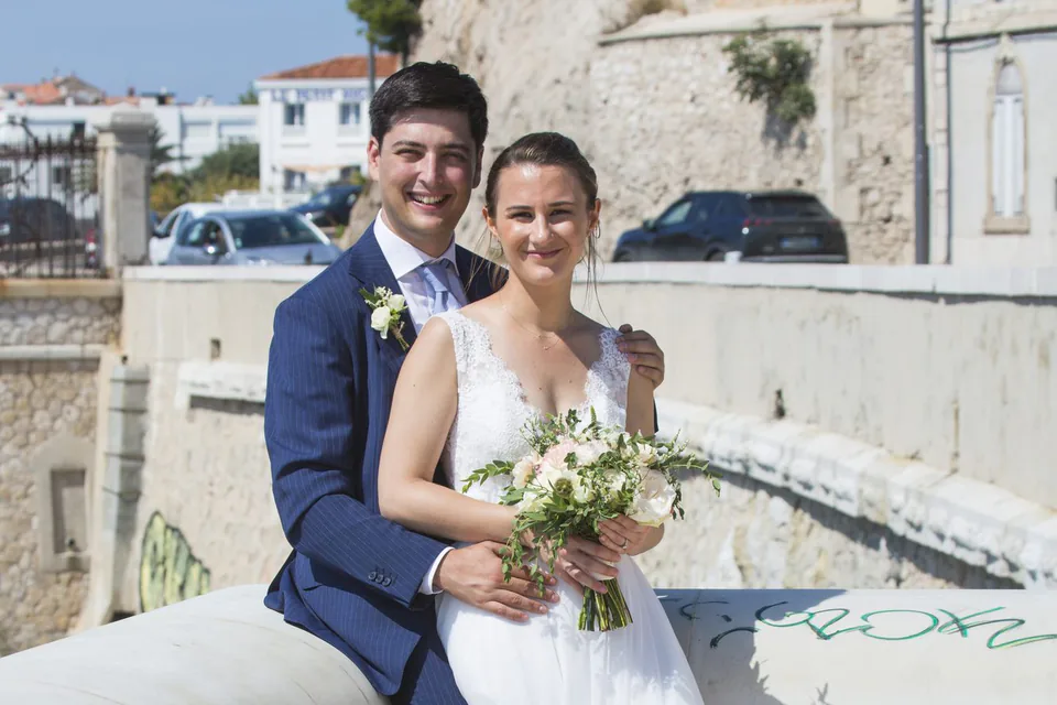 Couple de mariés assis sur un muret en ville, la mer en arrière-plan, à Marseille