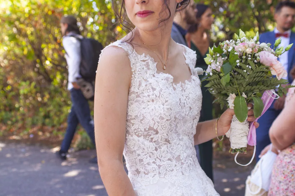Mariée avec son bouquet dans la lumière, invités flous en arrière-plan, à la sortie d’une cérémonie à Marseille